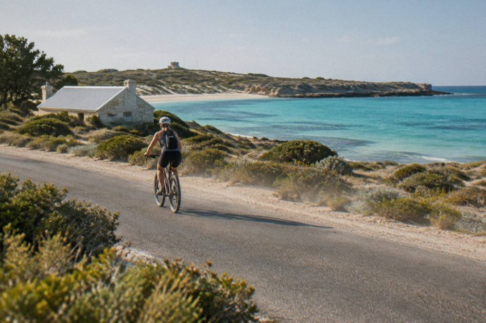 A cyclist on a coastal path beside calm turquoise water with low scrub and dunes