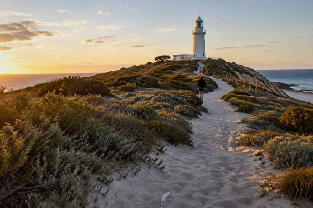 A sandy walking trail leading through low coastal heath toward a white lighthouse on a low hill