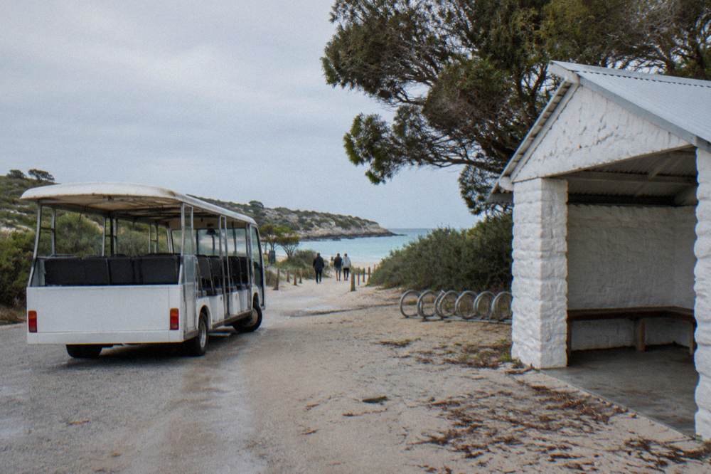 A hop-on hop-off bus parked beside a low stone building at a seaside settlement with bikes leaning on a rack