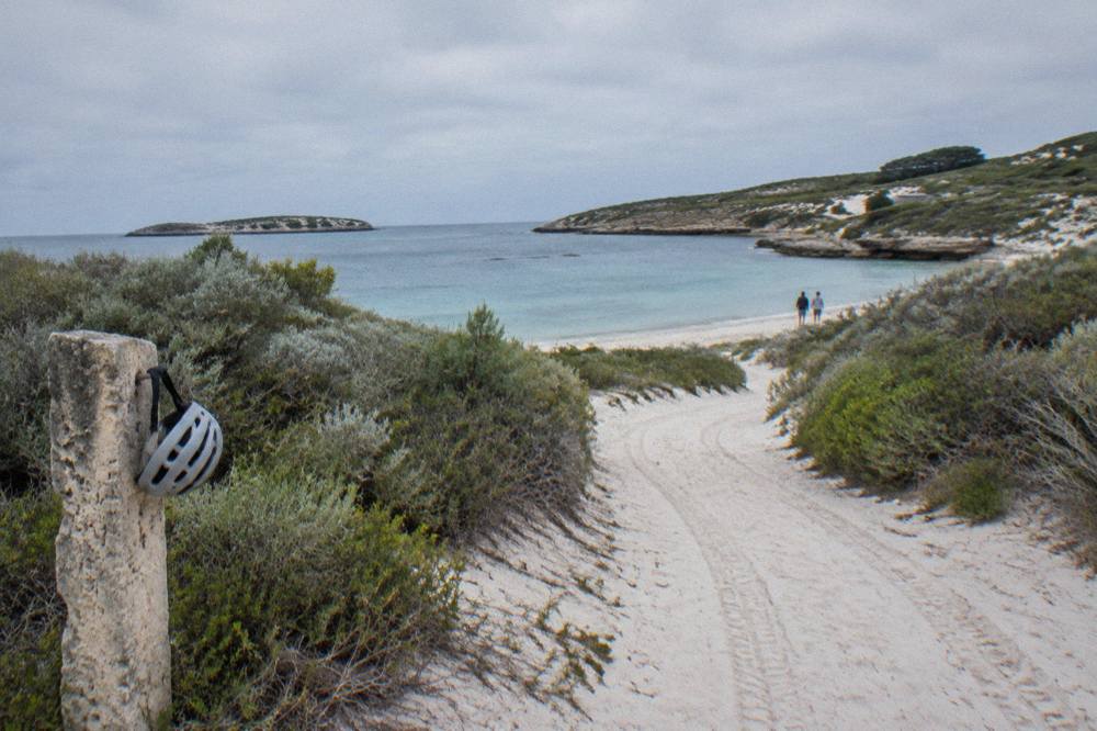 Bicycles parked on a sandy path beside low coastal scrub near a Rottnest bay