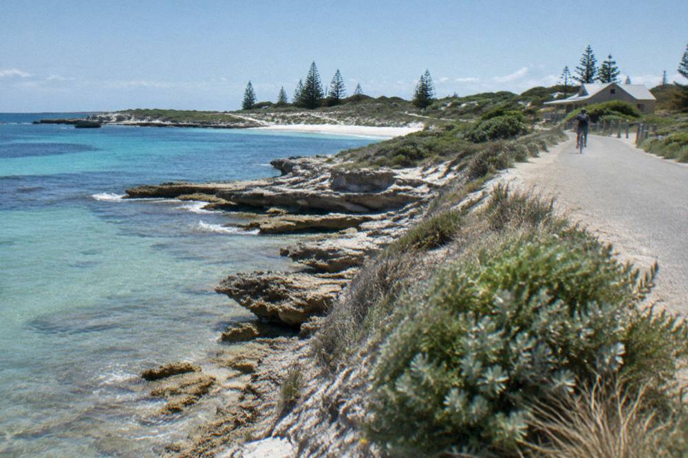 A cyclist riding a path above a rocky shoreline with clear shallow water below