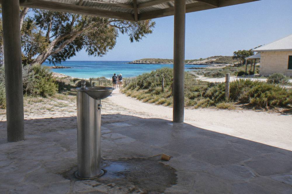 A shaded drinking fountain beside a sandy path with bicycles leaning against a fence near the Thomson Bay settlement.