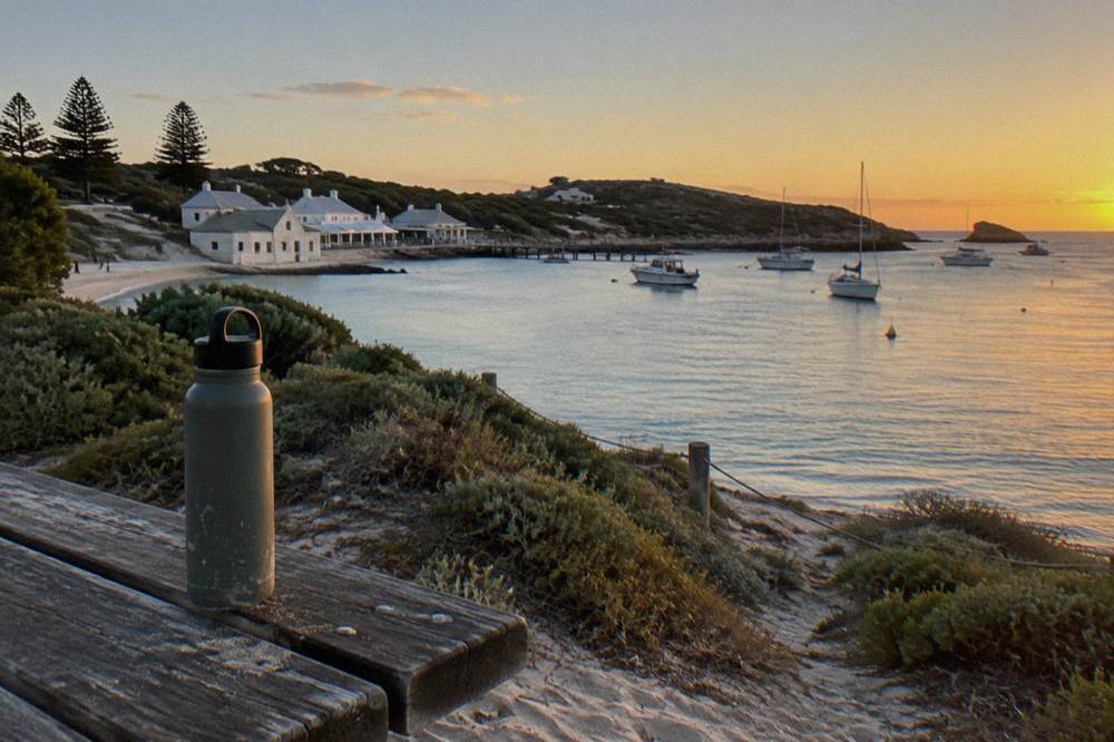 A reusable water bottle standing on a wooden bench overlooking the beach and moored boats at Thomson Bay on Rottnest Island.