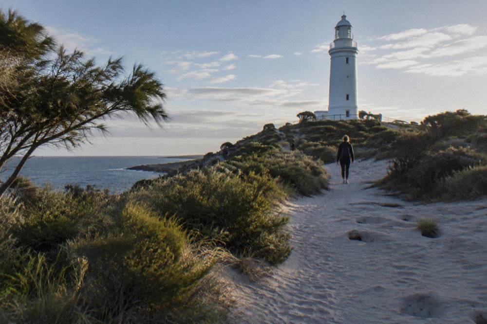A quiet sandy path between coastal scrub leading toward a lighthouse on Rottnest Island