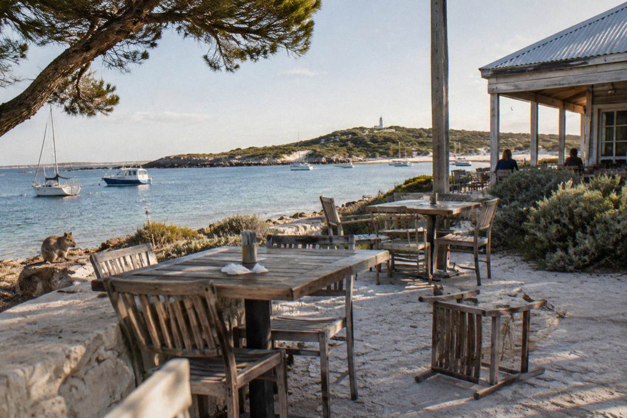 Outdoor tables beside a weathered timber pub building overlooking a calm bay with moored boats