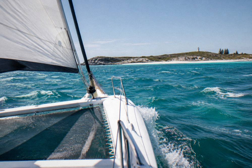 A catamaran under sail on open water with an island coastline on the horizon