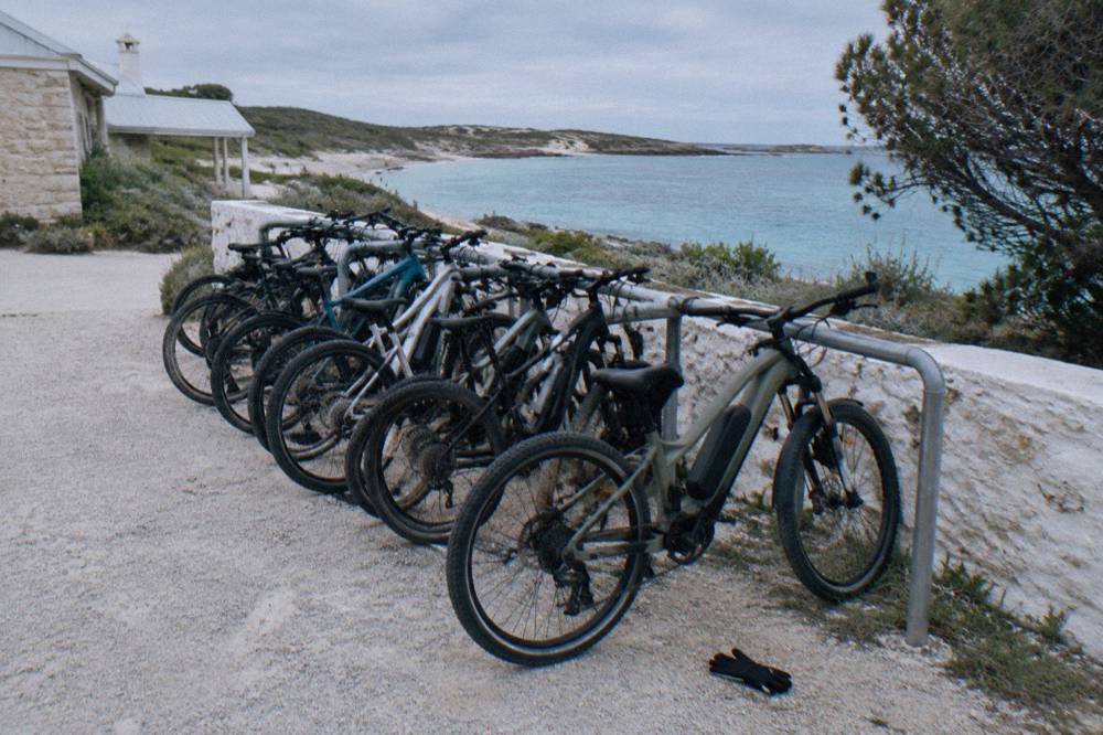 A row of rental bicycles locked to a rack near a low seaside building