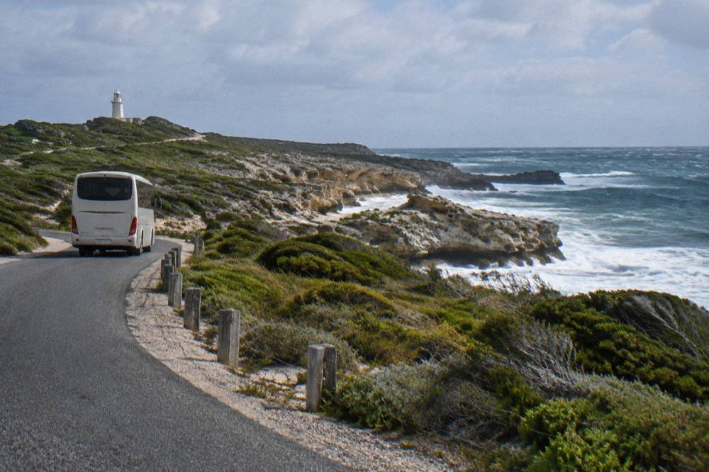 A tour bus winding along a narrow road between low coastal scrub and a blue bay