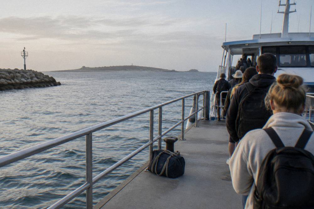 Passengers boarding a ferry at a mainland terminal jetty on a calm morning with the island visible on the horizon