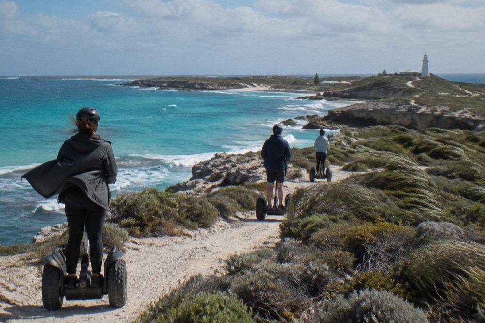 Segway riders moving along a sandy coastal track above a calm turquoise bay