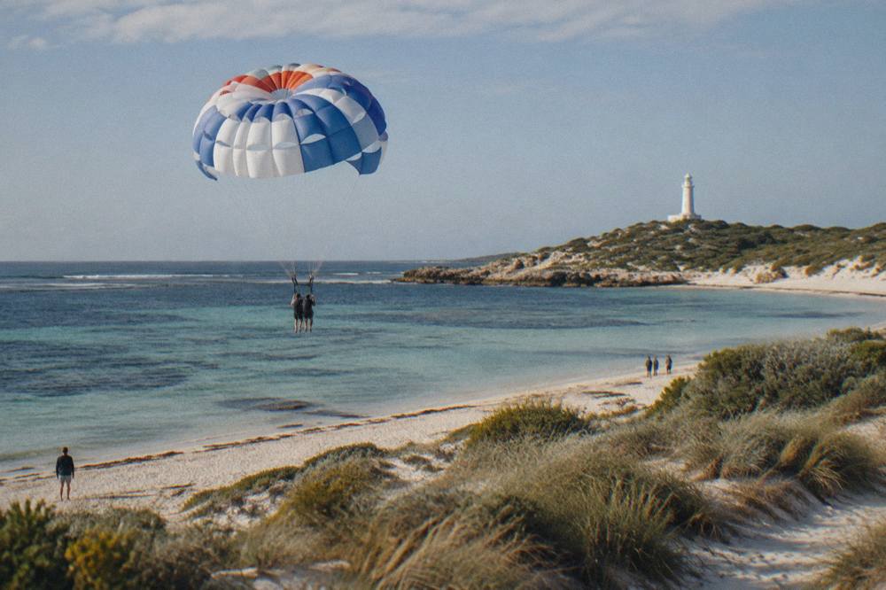 A tandem parachute canopy descending toward a pale sandy beach with clear shallow water